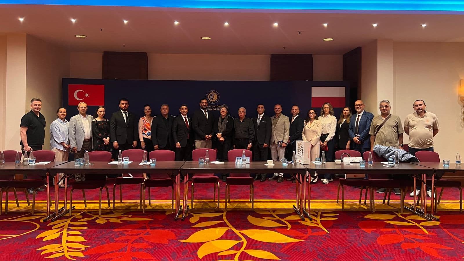 About two dozen participants standing behind a long boardroom table at TURKPOL’s market information meeting in Warsaw, with Turkish and Polish flags behind them.